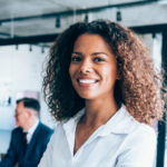 A woman in a smart shirt, standing in an office with colleagues, smiling.