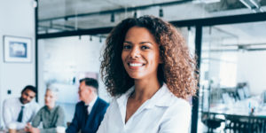 A woman in a smart shirt, standing in an office with colleagues, smiling.