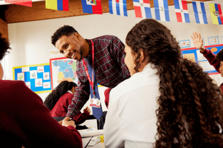 Teacher wearing shirt, tie and lanyard leaning over desk to help teenage student with work