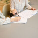 Woman signing on white paper while sat next to another woman with printed work documents. Both women are in work attire.